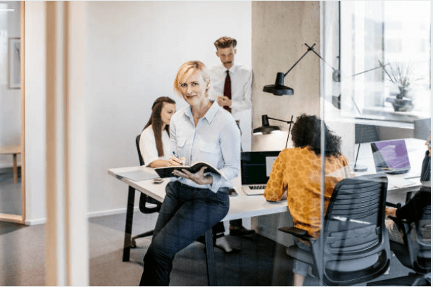 A professional woman holding a notebook, standing confidently in a collaborative office environment with colleagues working in the background.