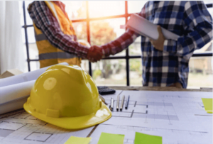A yellow hard hat on a table with construction blueprints, as two people shake hands in the background, symbolizing a construction agreement or partnership.