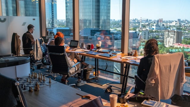 Employees working at desks in a modern office with large windows overlooking a cityscape.