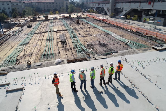 A group of construction workers wearing safety gear standing on a large building site with visible rebar and materials.